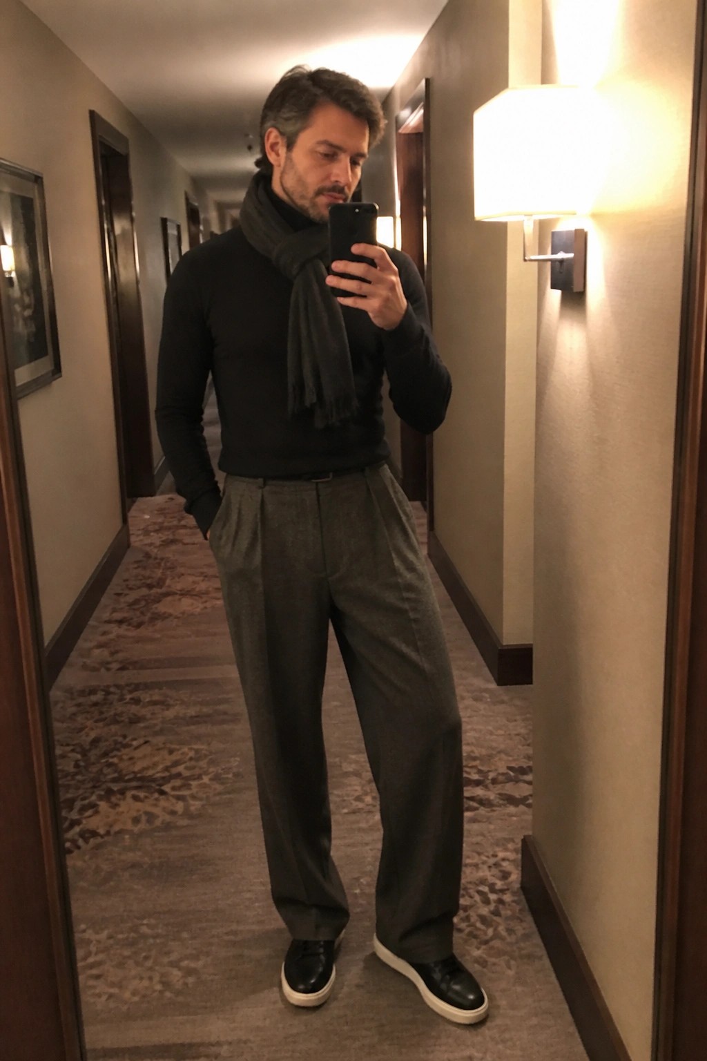 Man in black turtleneck and scarf taking mirror selfie in hallway wearing gray pants