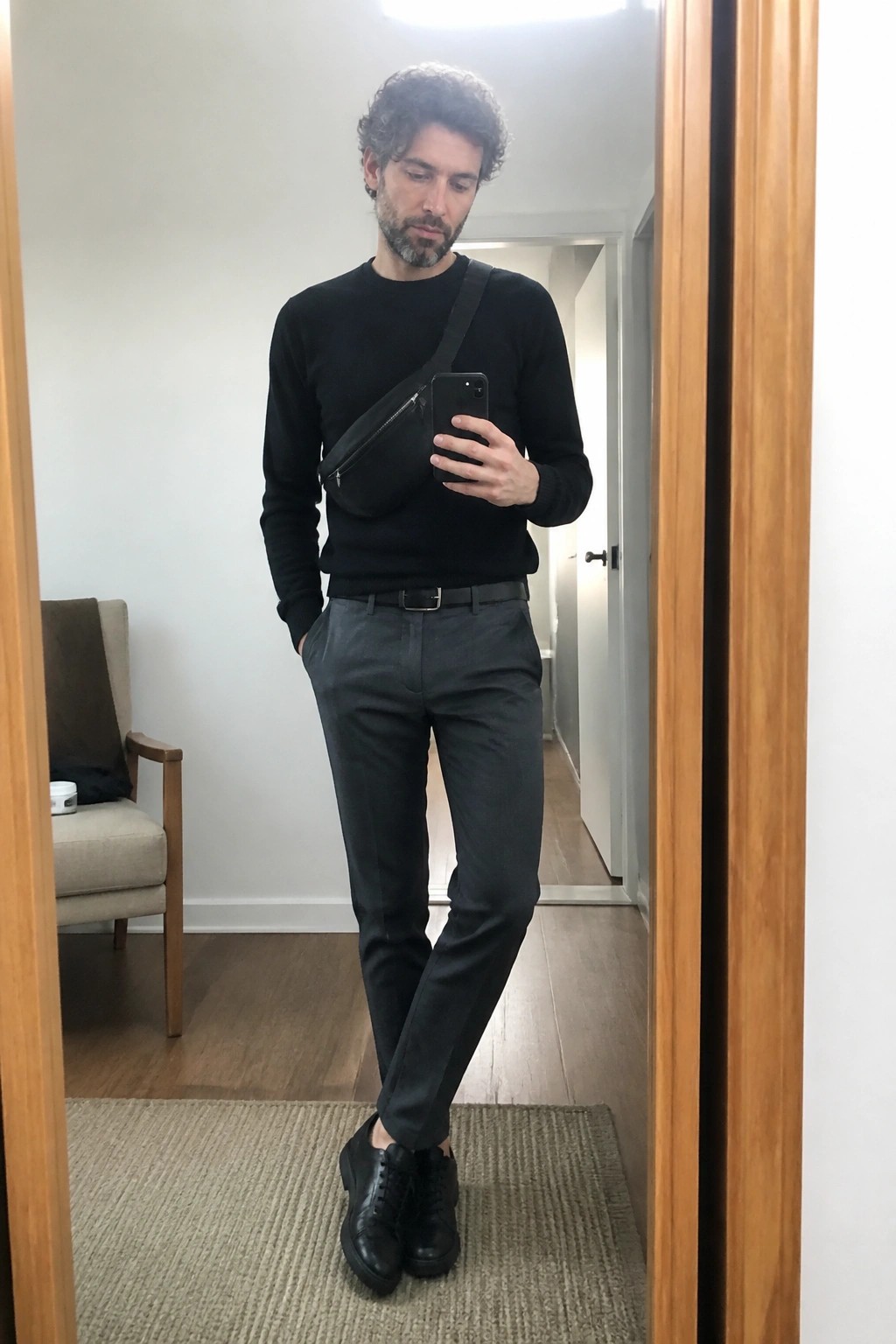 Man with curly hair taking mirror selfie in black sweater and gray pants with bag