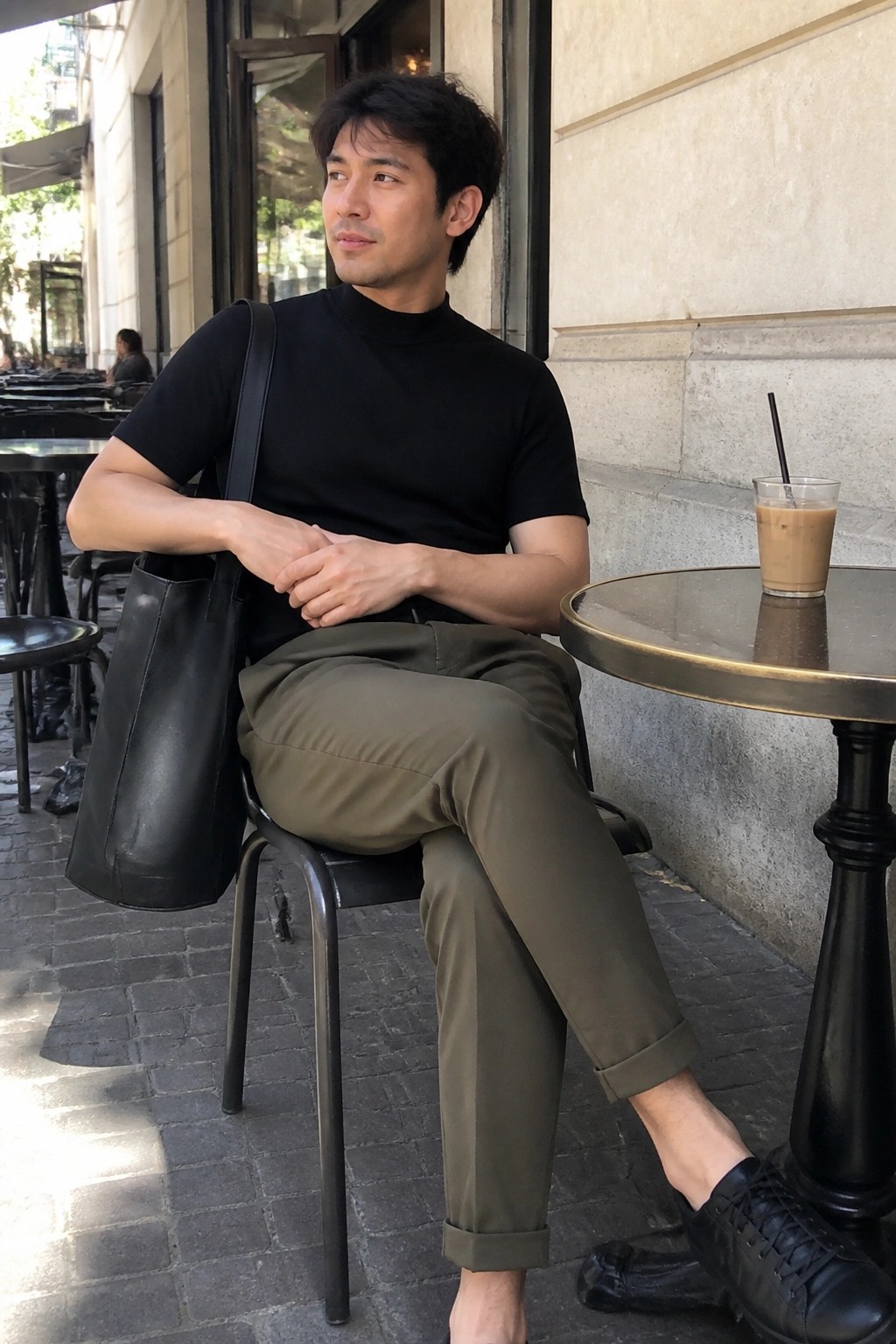 Man in black turtleneck and olive pants seated at cafe with iced coffee
