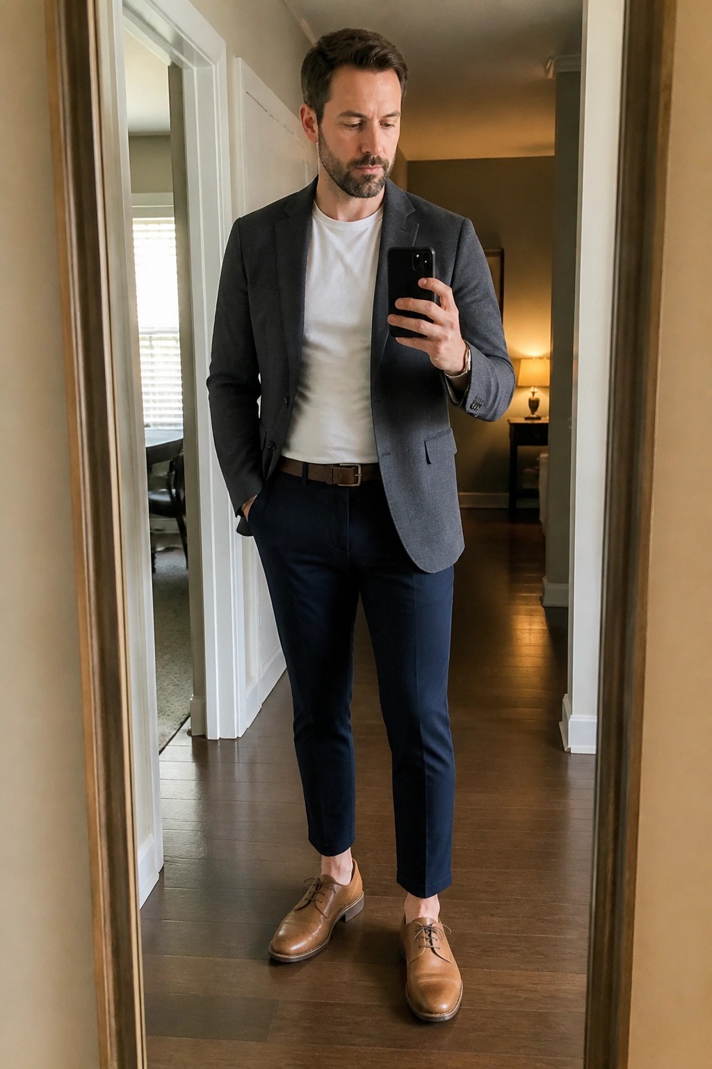 Man in gray blazer and navy pants taking mirror selfie with phone.