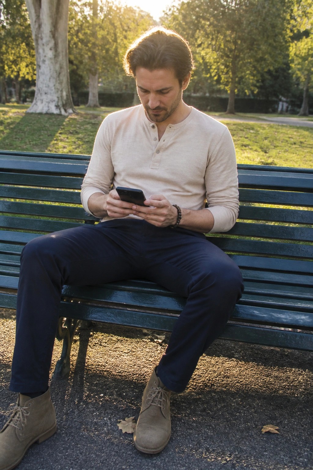 Man in beige shirt and navy pants uses smartphone on green park bench.