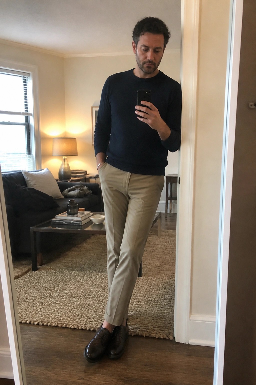 Man in navy sweater and khaki pants taking mirror selfie in living room