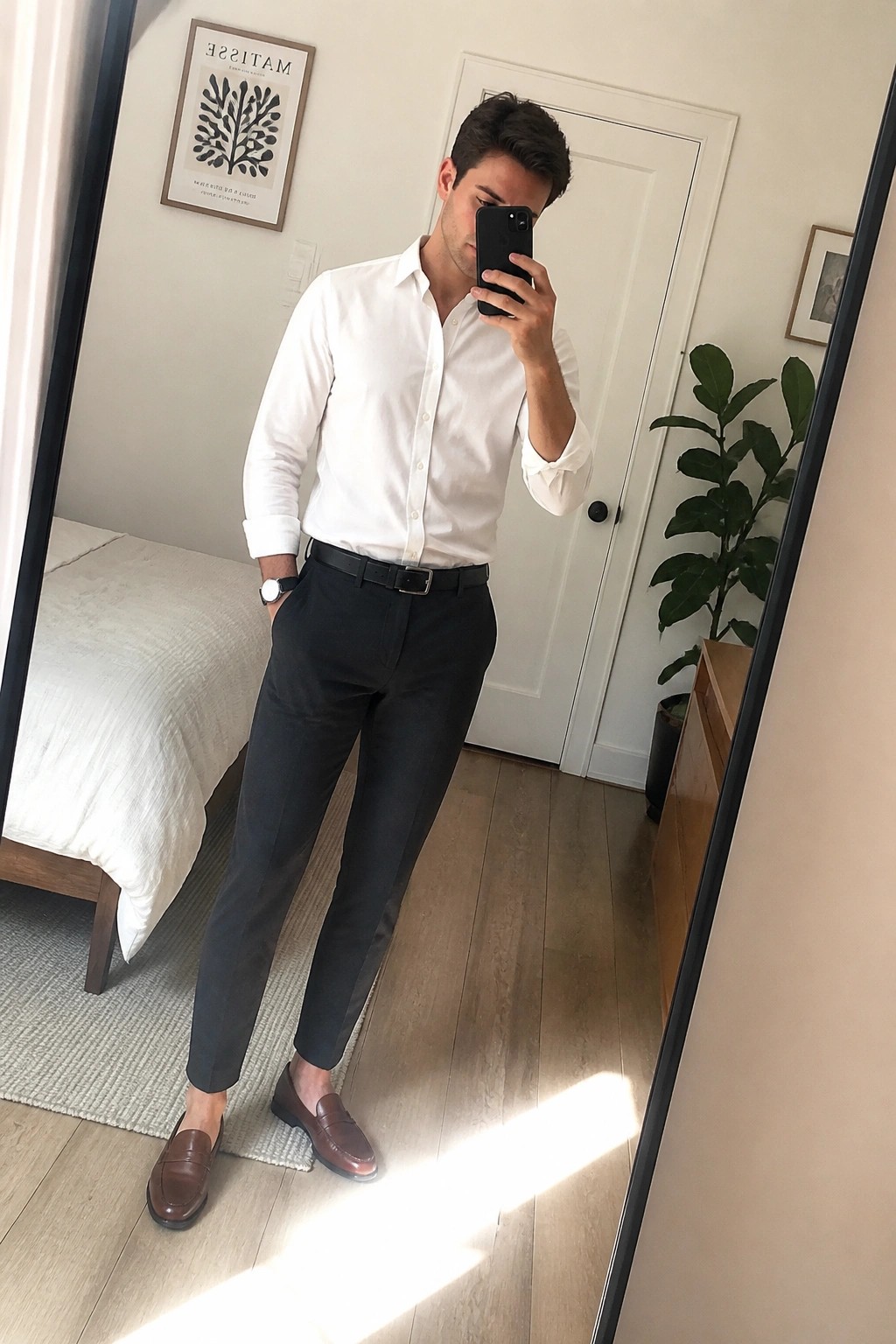 Man in white shirt and dark pants taking bedroom mirror selfie with phone.