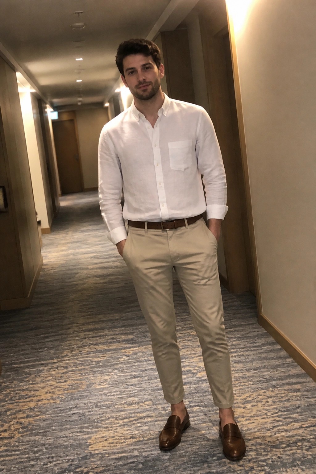 Man in white shirt and beige pants standing in hotel hallway with hands in pockets.