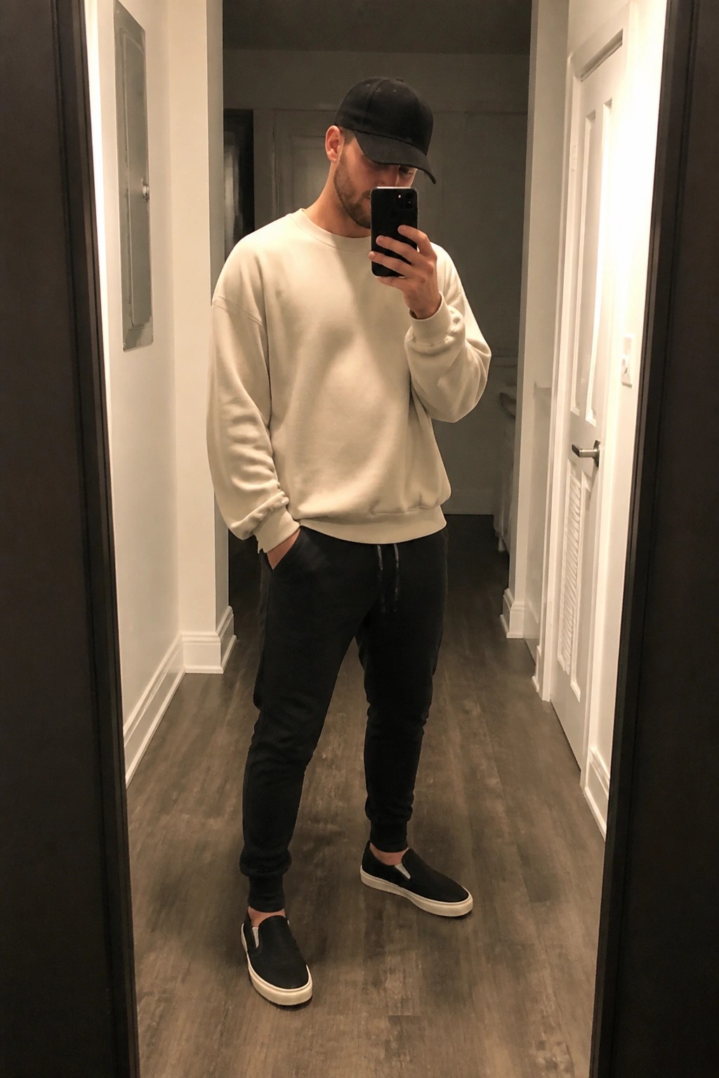 Man in cream sweatshirt and black cap taking mirror selfie in hallway