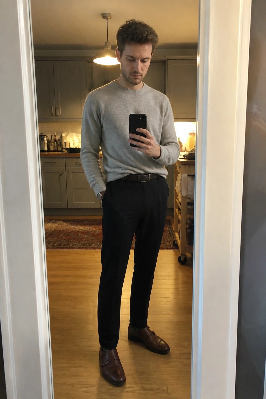 Man in gray sweater and black pants taking mirror selfie in kitchen