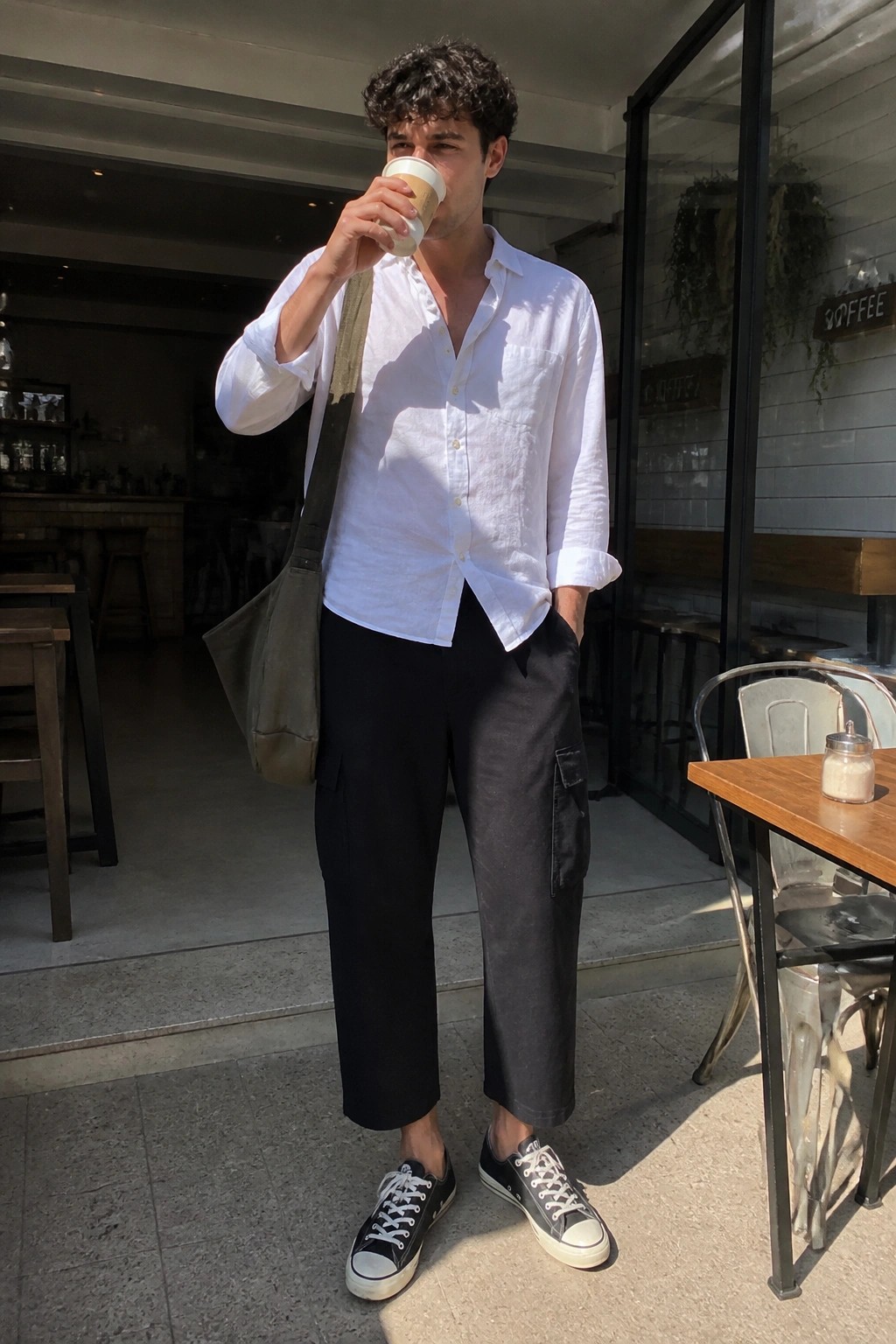 Man in white shirt and black cargo pants drinks coffee outside cafe.