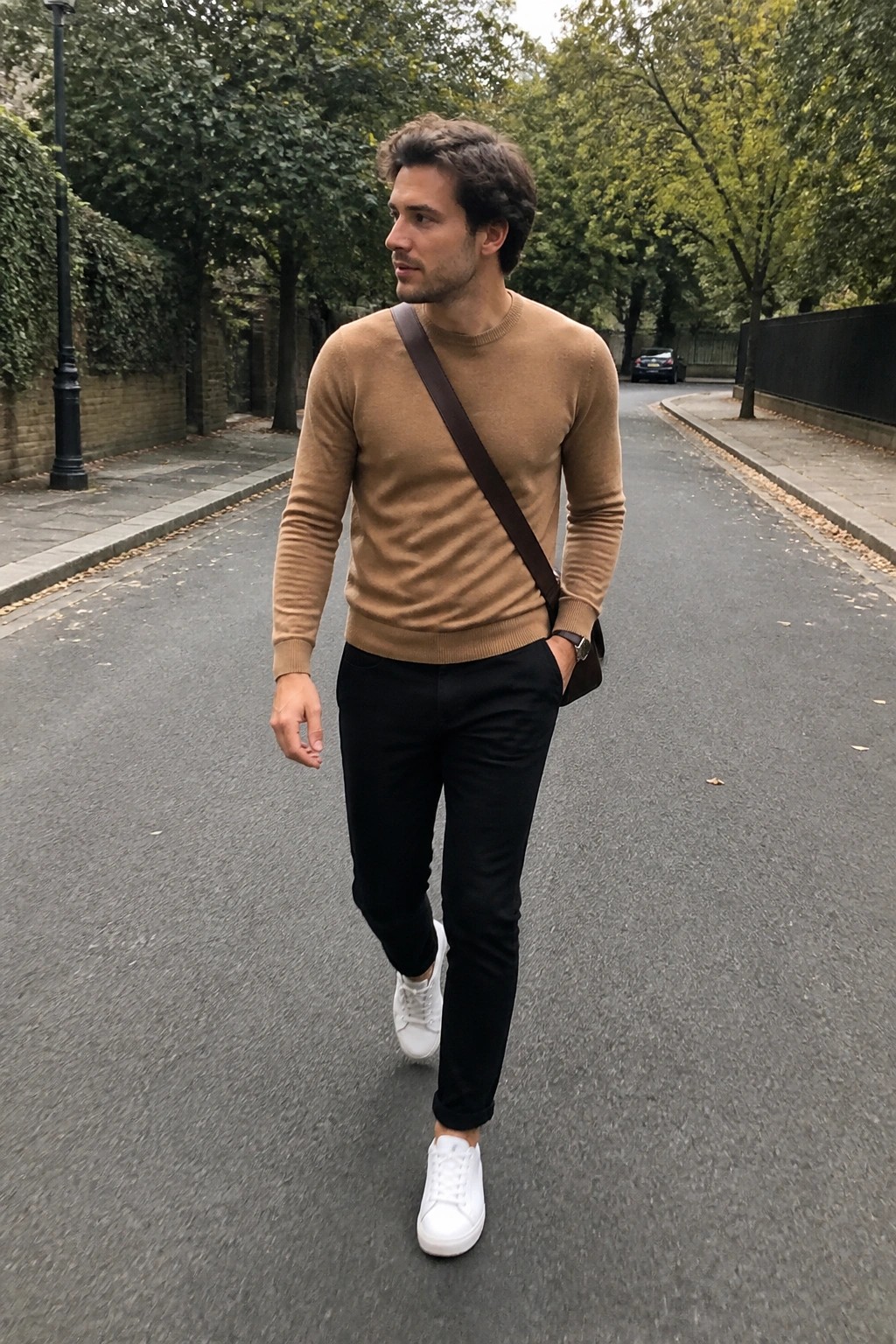 Man in tan sweater, black pants, white sneakers walks on leafy street with crossbody bag