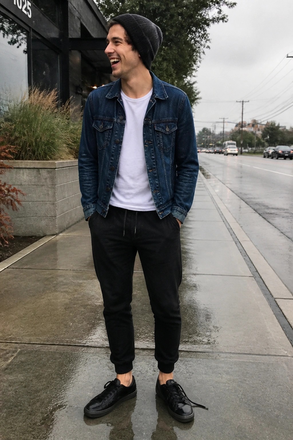 Man in denim jacket and beanie smiles on wet city sidewalk.