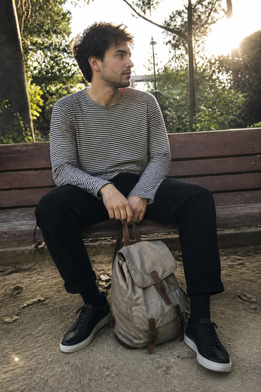 Man in striped shirt on wooden bench with beige backpack in sunlight