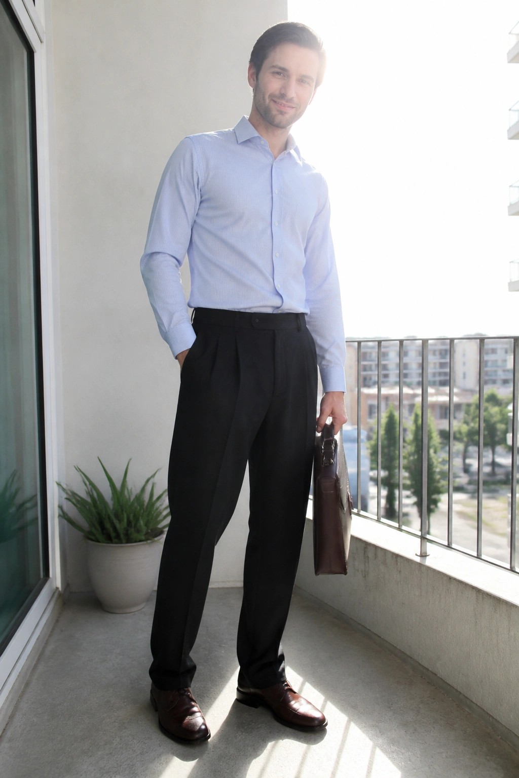 Man wearing light blue button down shirt tucked into black pleated trousers with brown leather dress shoes and holding a brown leather bag.