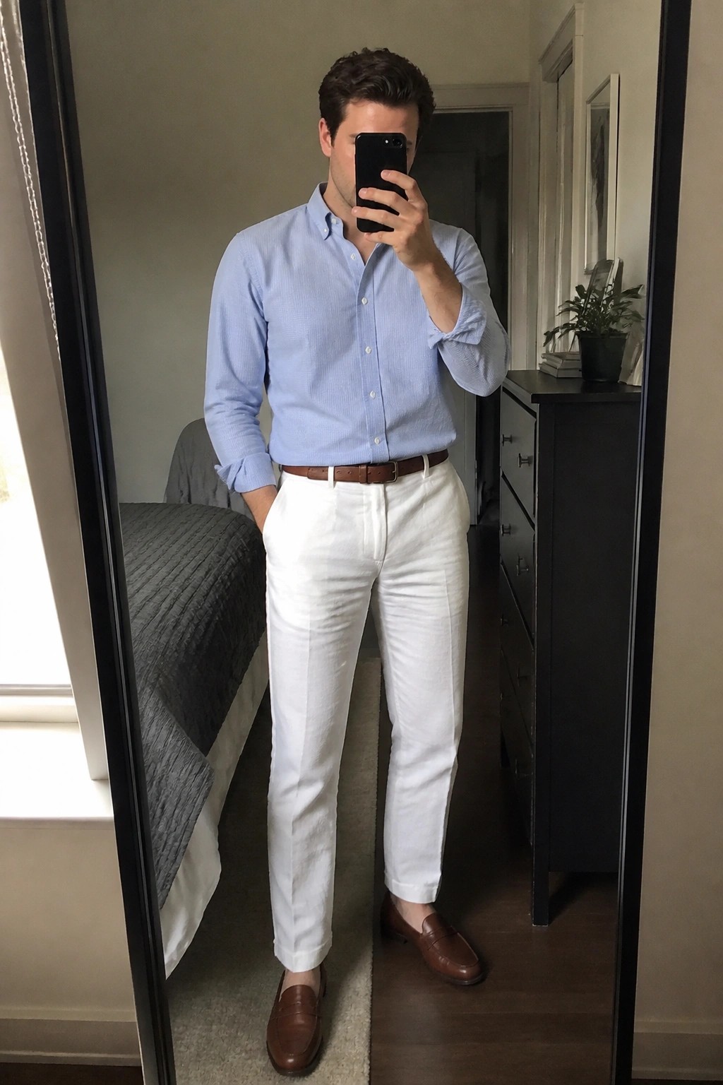Man in light blue shirt and white pants taking mirror selfie in bedroom