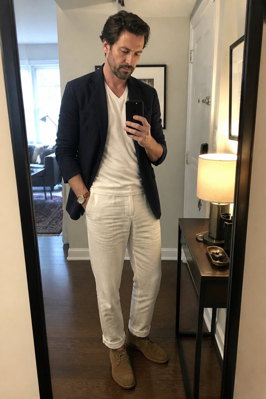 Man in navy blazer, white v-neck tee, white linen pants, and brown suede shoes taking a mirror selfie.