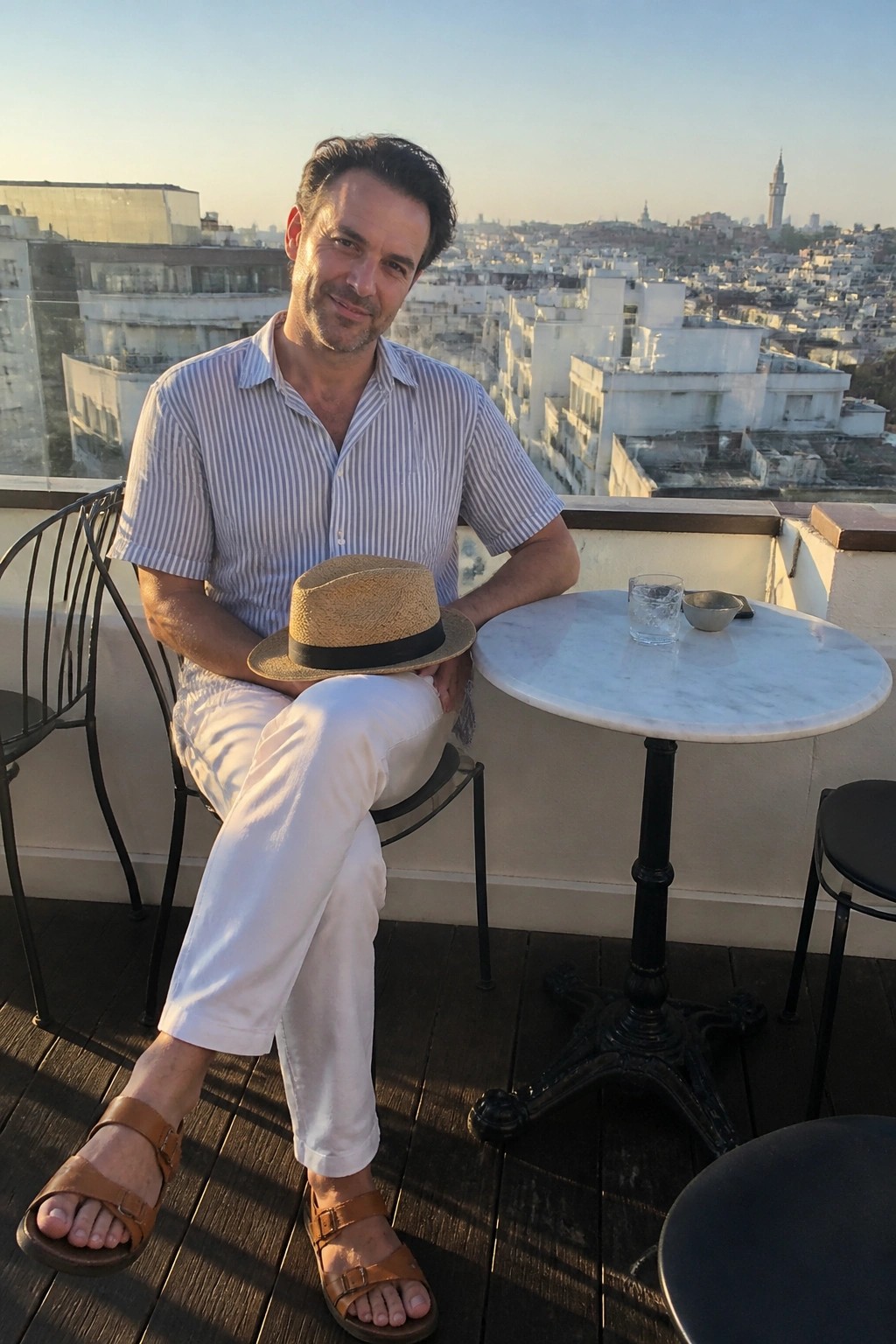 Man in striped shirt holding straw hat on sunny rooftop terrace overlooking city.