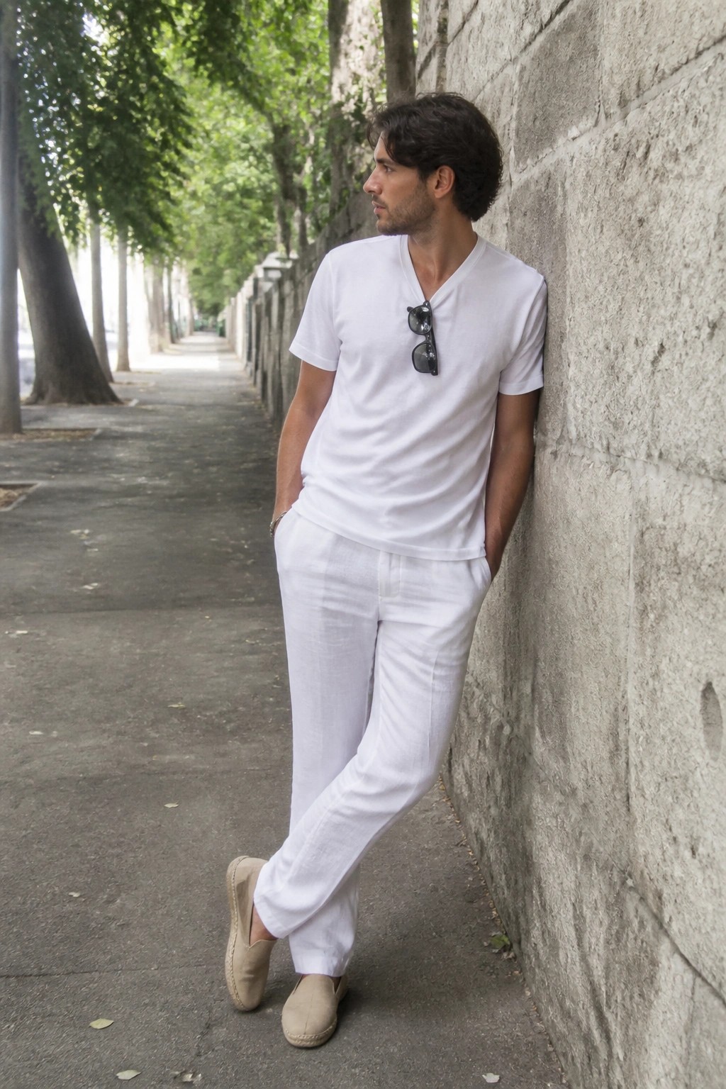Man in white shirt and pants leaning against stone wall on tree-lined path.