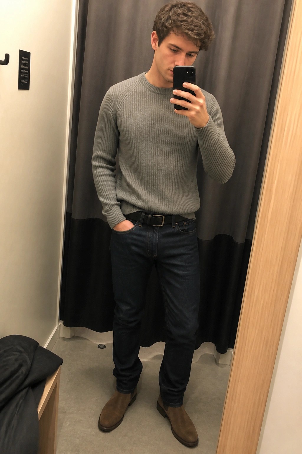 Man wearing gray ribbed sweater tucked into dark straight leg jeans with black belt and brown suede Chelsea boots while taking a mirror selfie in a fitting room.