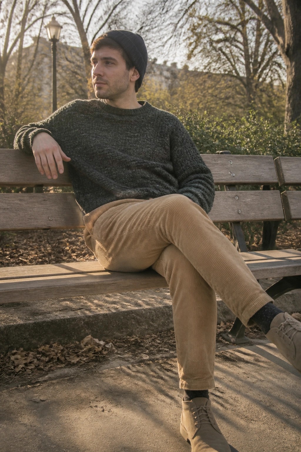 Man in beanie and corduroy pants lounges on sunny park bench.