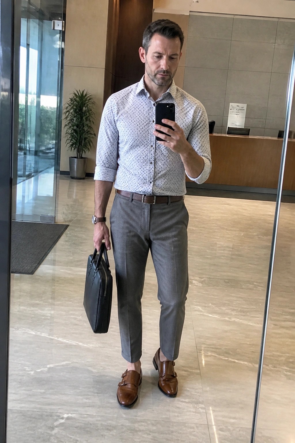 Man in patterned shirt and gray pants taking mirror selfie with briefcase in lobby