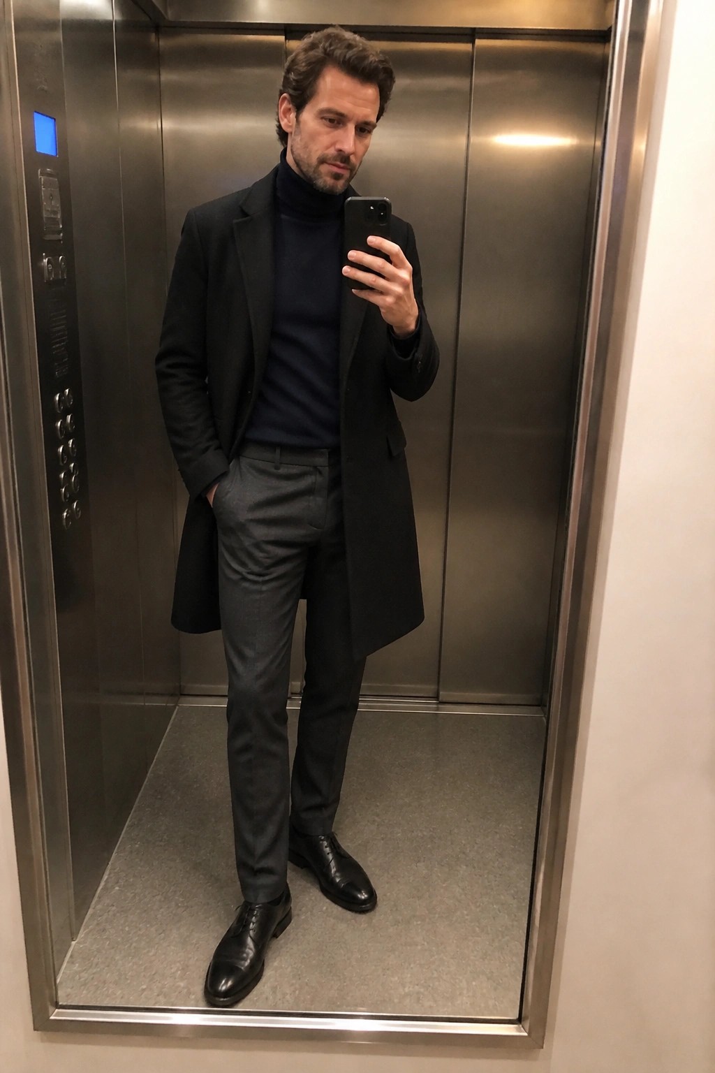 Man in black overcoat taking mirror selfie inside metal elevator