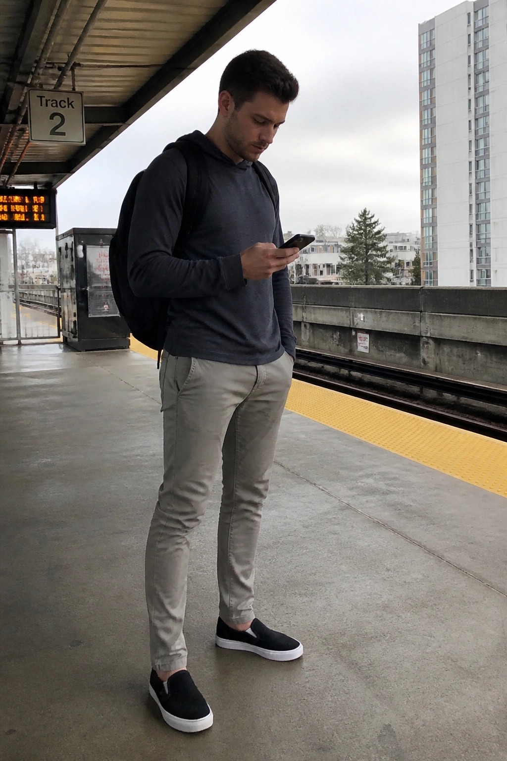 Man in gray hoodie and backpack checks phone on train platform