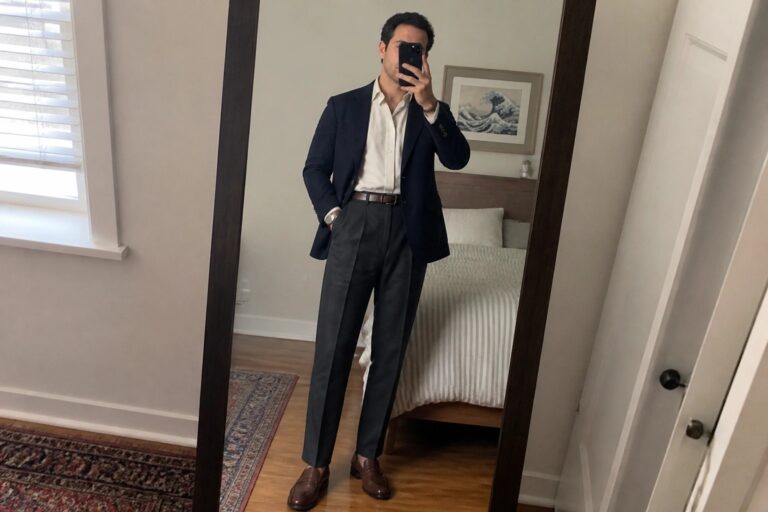 Man in navy blazer and gray trousers taking bedroom mirror selfie.