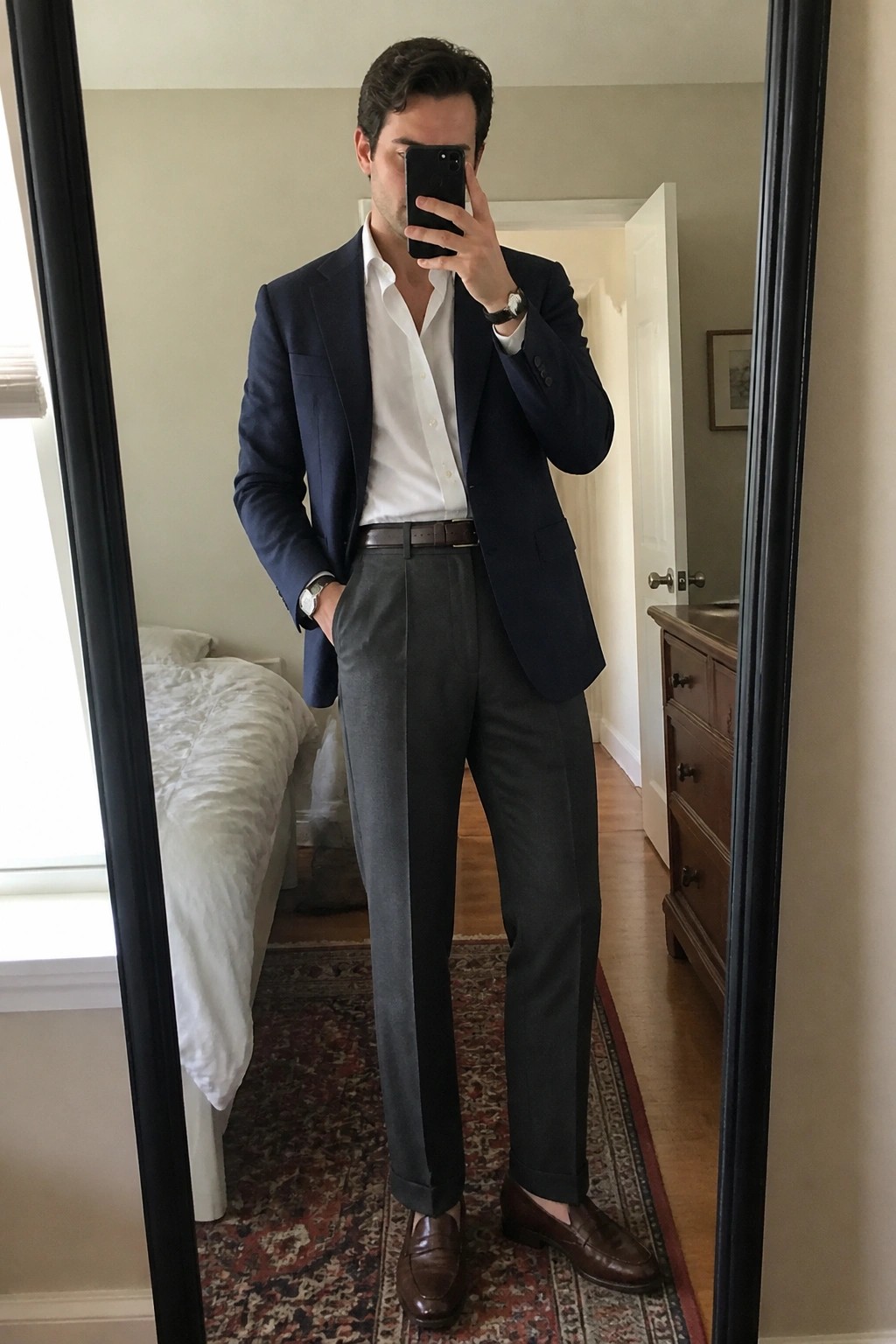 Man in navy blazer and gray trousers taking bedroom mirror selfie.