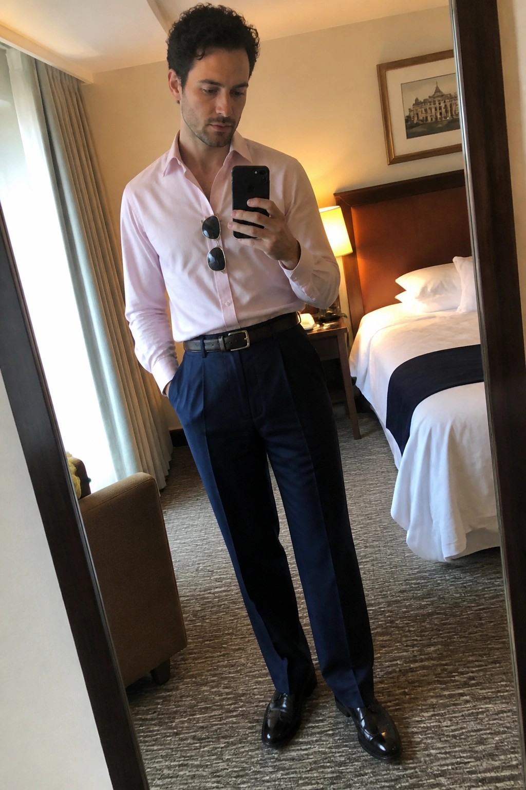 Man in pink shirt and navy pants taking mirror selfie in hotel room