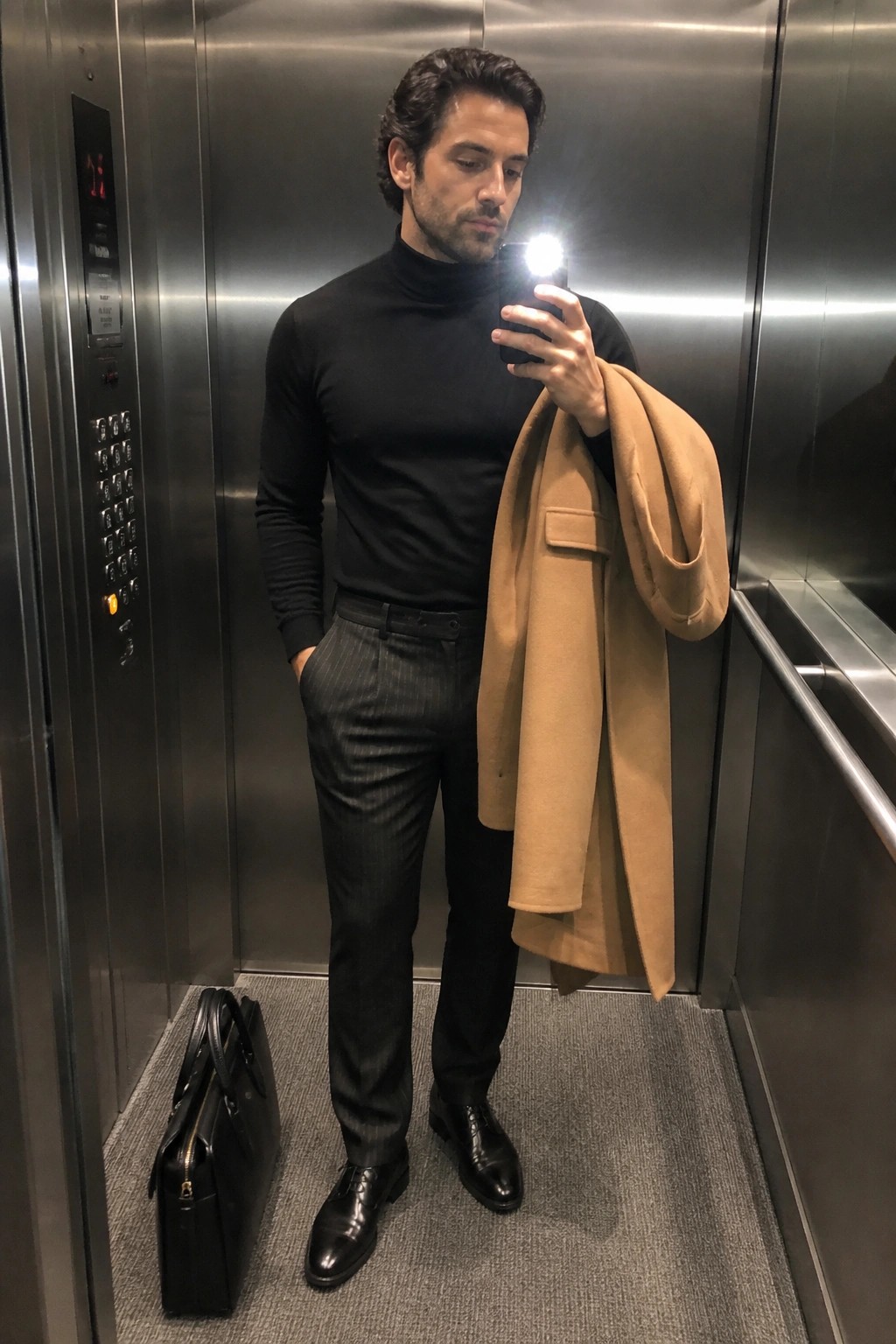 Man in black turtleneck taking elevator selfie, holding tan coat with briefcase nearby.