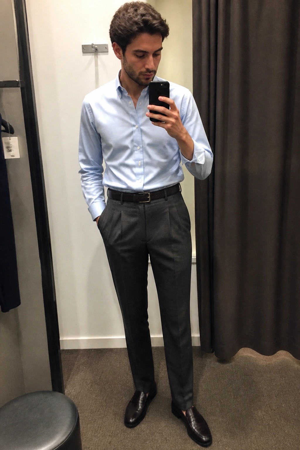 Man in light blue shirt and gray trousers taking mirror selfie in fitting room