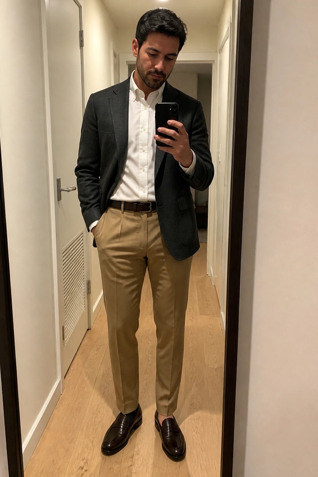 Man in dark gray blazer over white button down shirt with pleated khaki trousers brown belt and matching loafers taking a mirror selfie in a hallway.