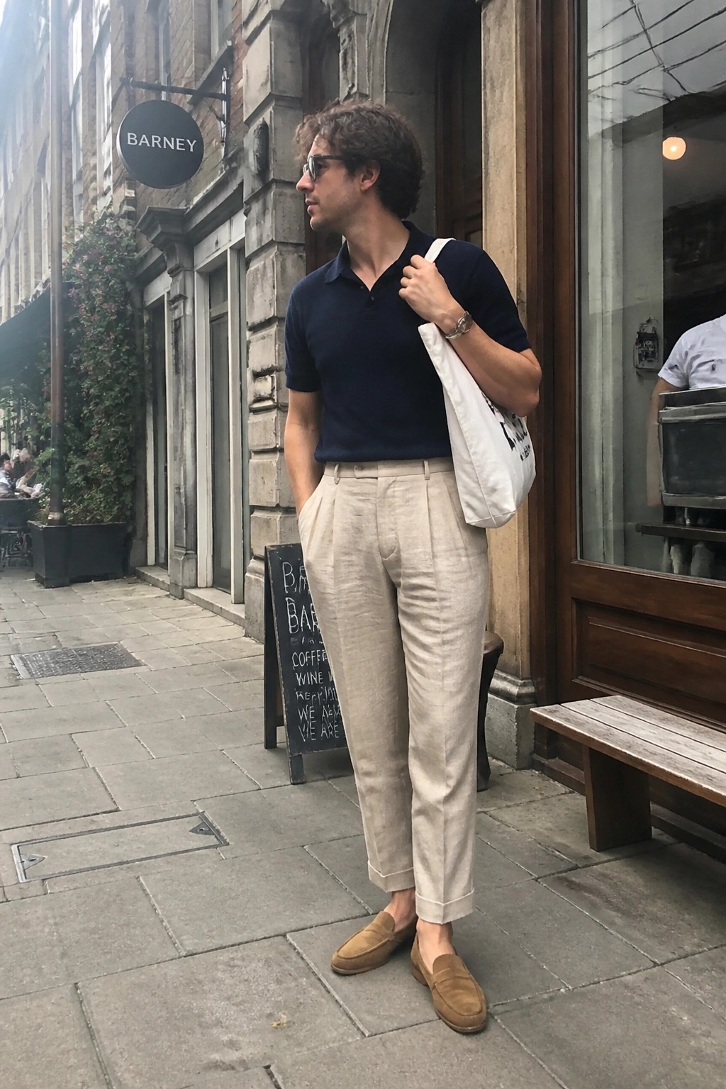 Man in navy polo and beige linen pants with white tote on city sidewalk.