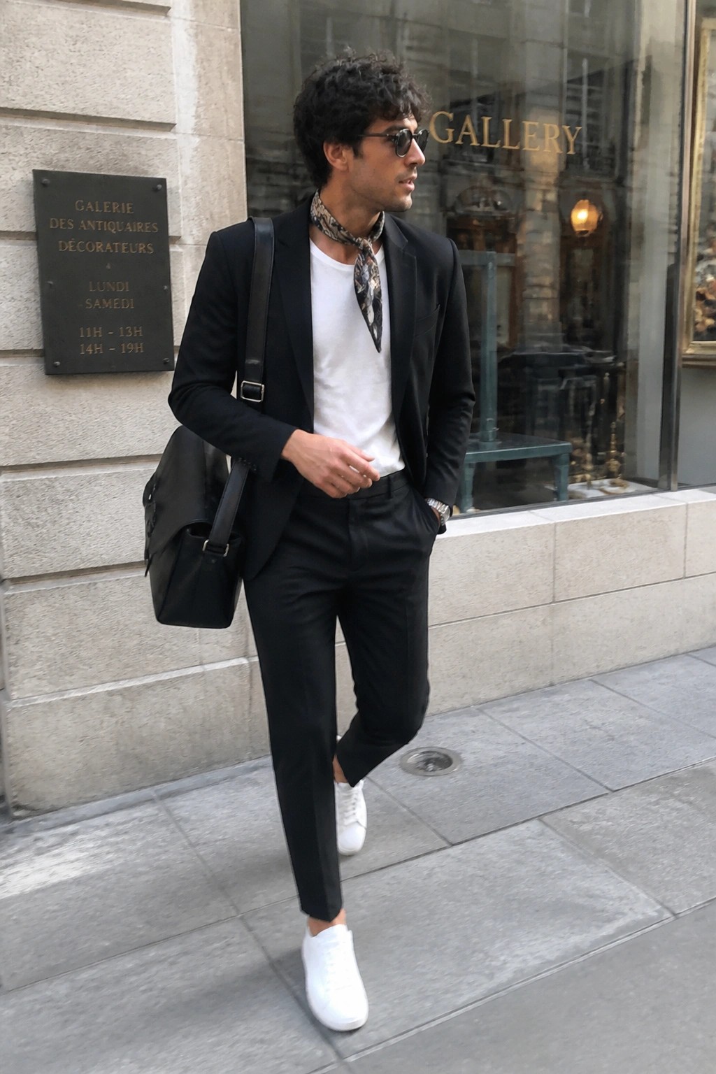 Man in black blazer, white tee, patterned scarf, and white sneakers with shoulder bag on sidewalk