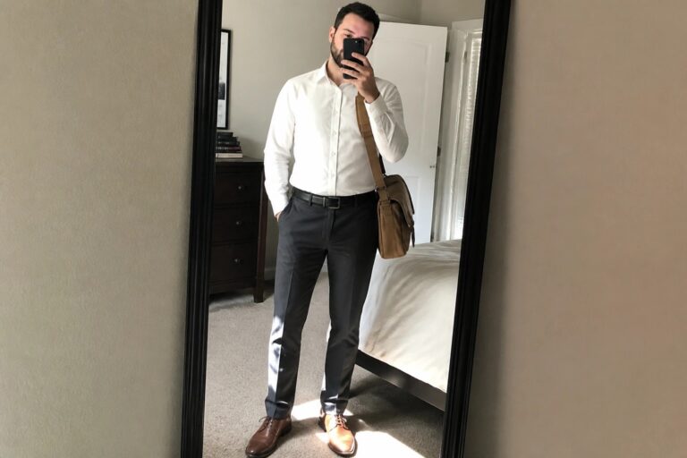 Man in white shirt and dark pants taking mirror selfie in bedroom