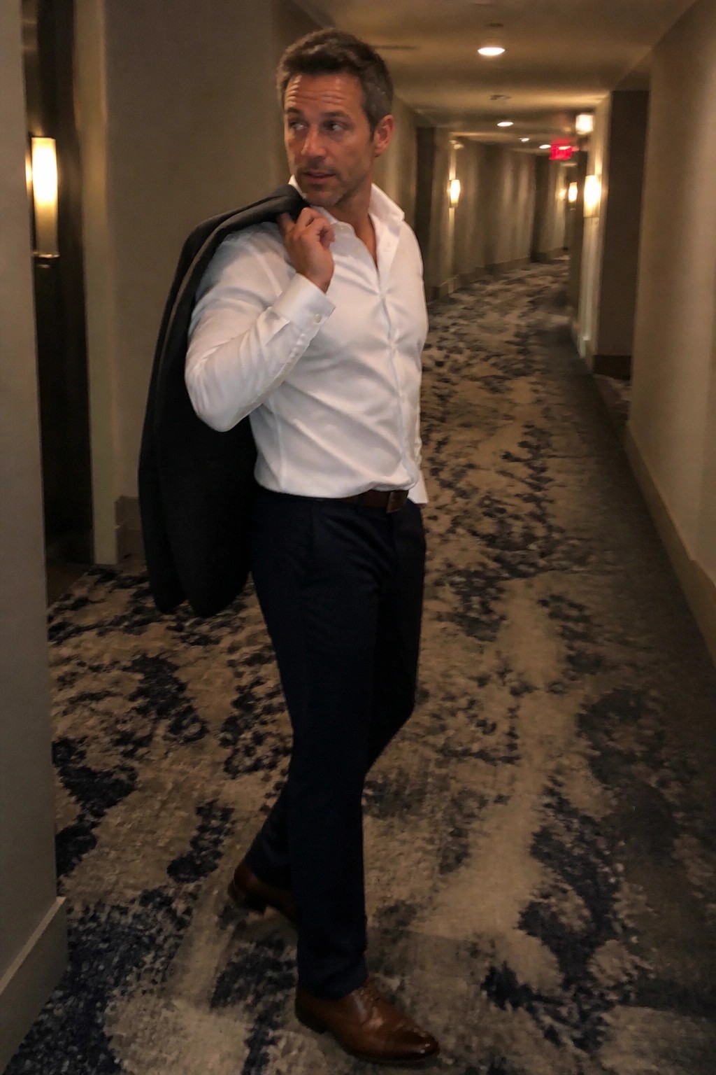 Man in white shirt holding black jacket over shoulder in hotel hallway