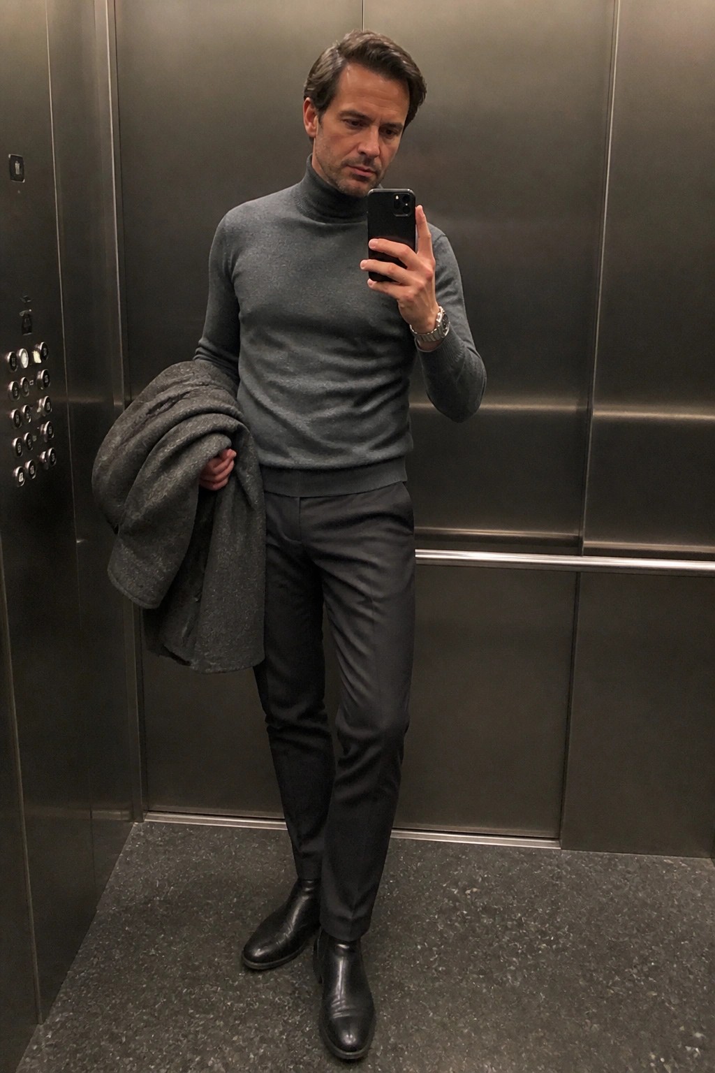Man in gray turtleneck holding phone and coat in metal elevator