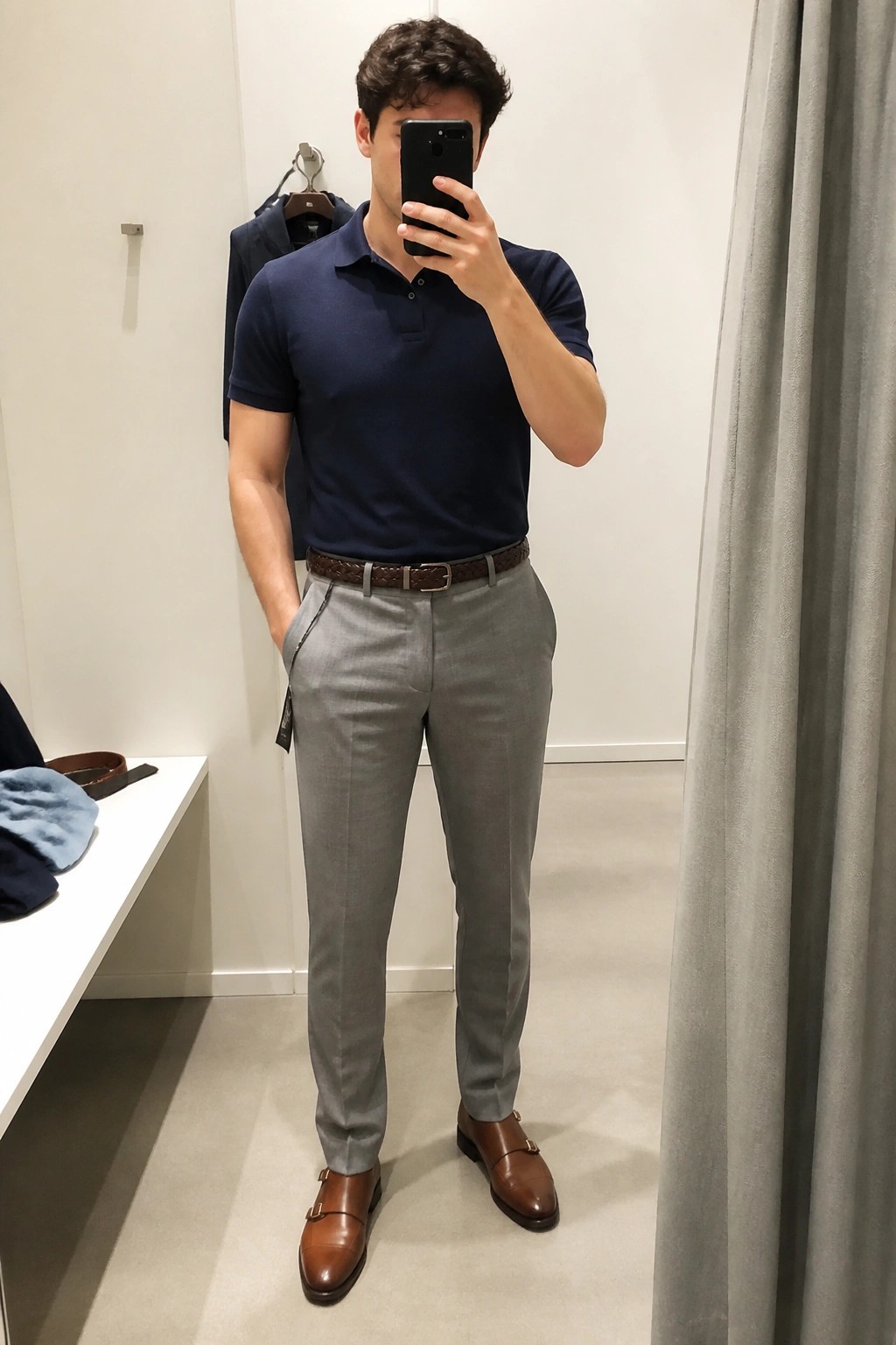 Man in navy polo shirt and gray pants taking mirror selfie in fitting room.