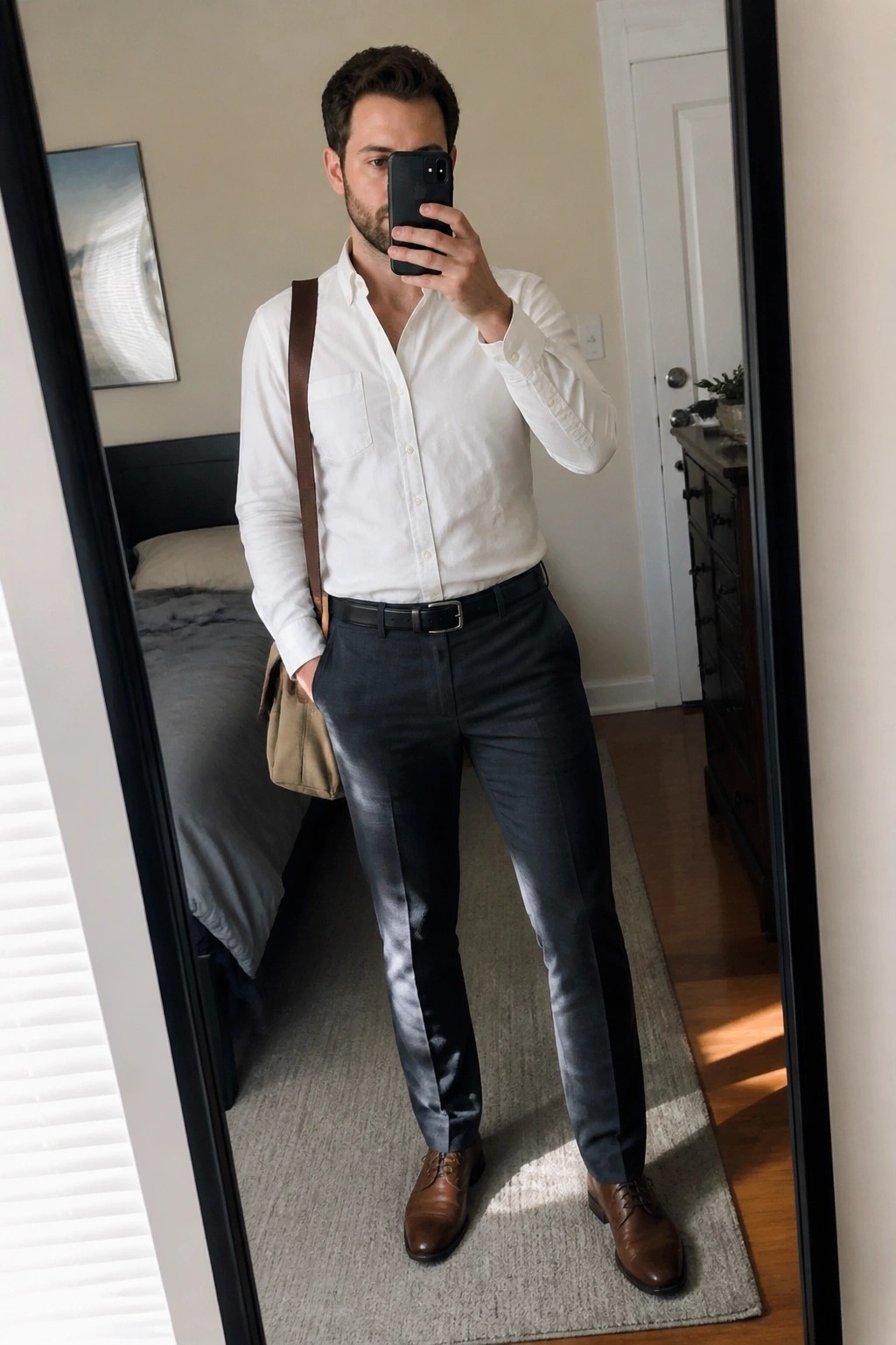 Man in white shirt and dark pants taking mirror selfie in bedroom