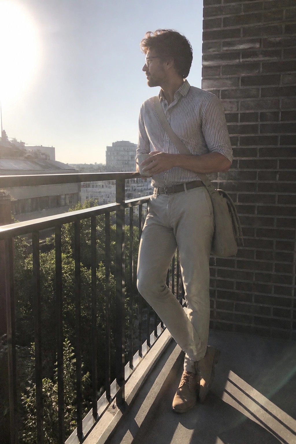Man in striped shirt and beige pants holding cup on sunny city balcony