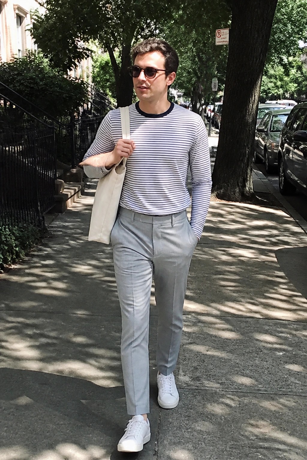 Man in striped shirt and gray pants with tote bag on sunny sidewalk
