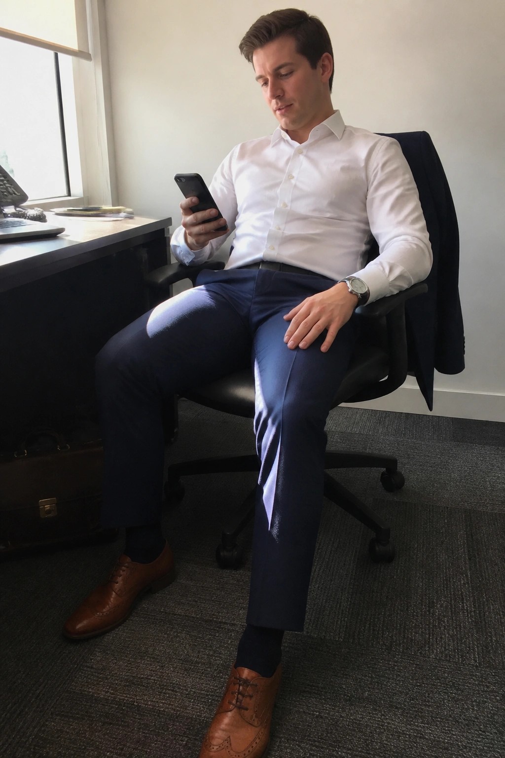 Young man in white shirt and blue pants checks smartphone in office chair