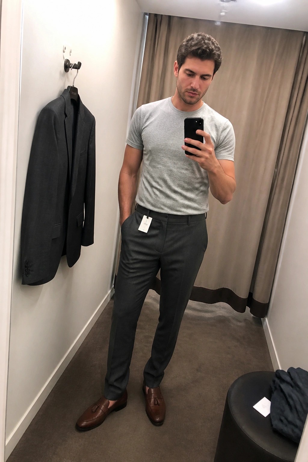 Man in gray t-shirt and pants taking mirror selfie in fitting room with hanging blazer