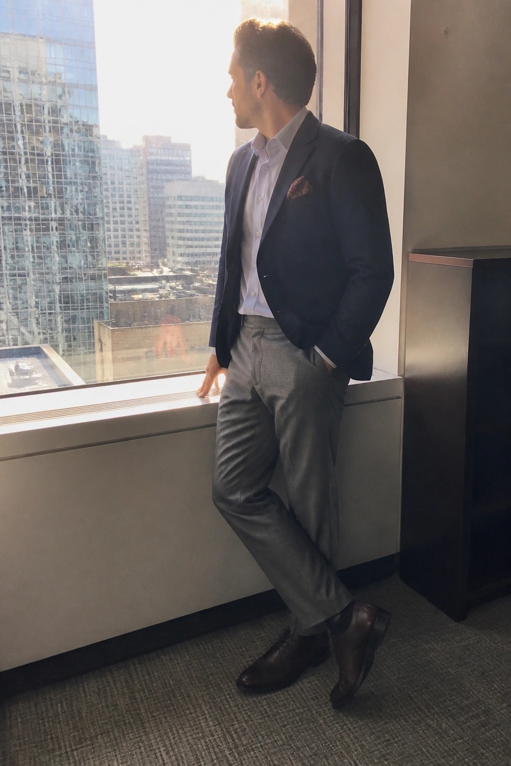 Man in navy blazer and gray pants by sunny office window overlooking cityscape