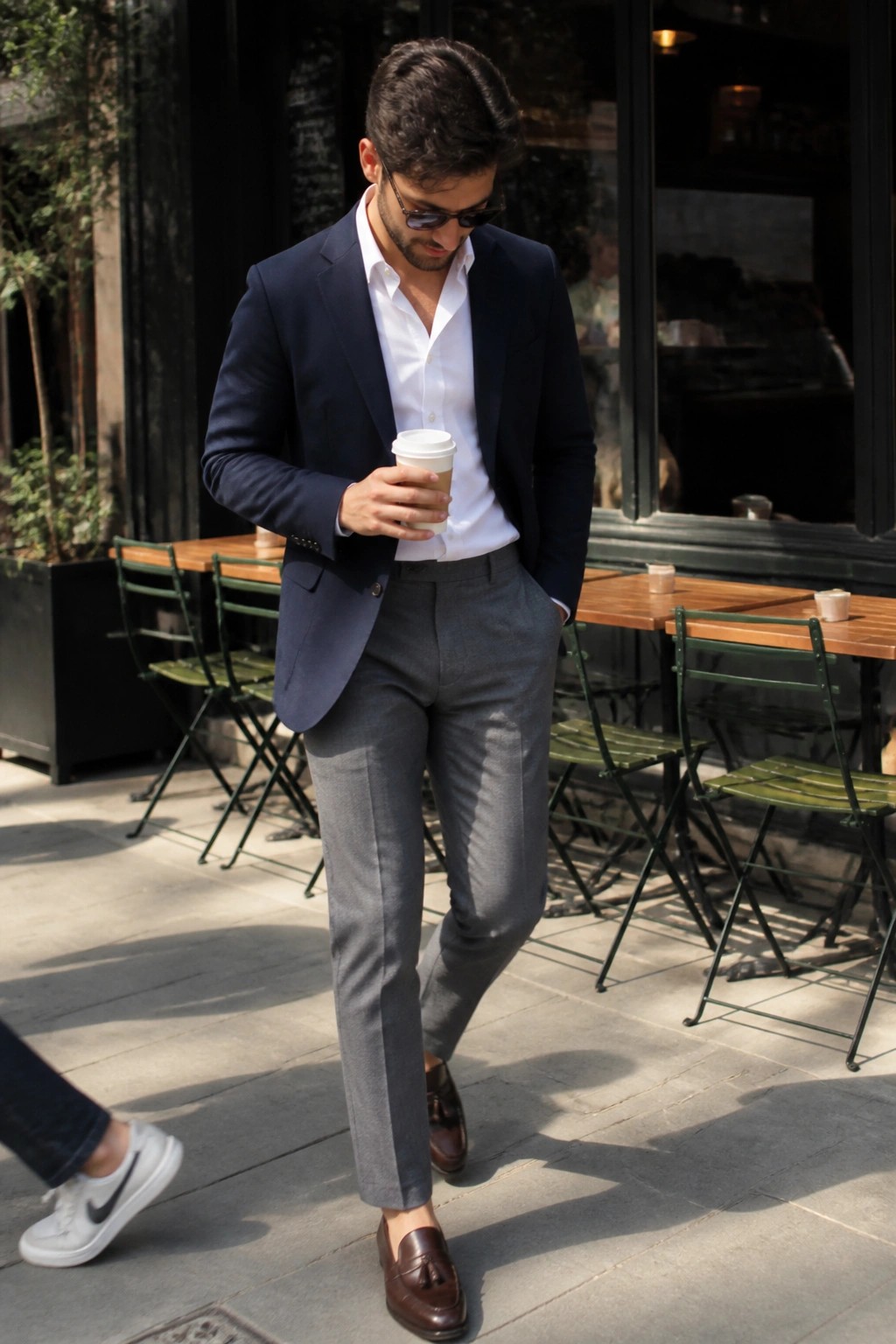 Man wearing navy blazer over white shirt with grey formal trousers and brown tassel loafers while holding a coffee cup outdoors