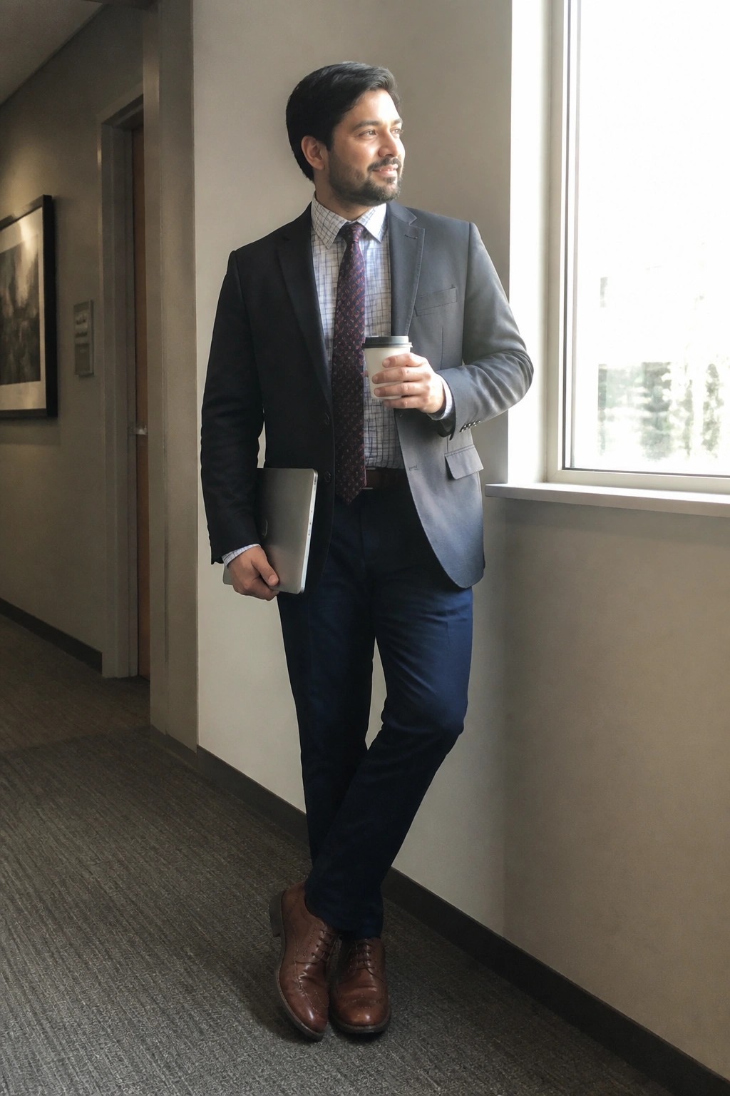 Man in gray blazer and blue pants holds laptop and coffee by window