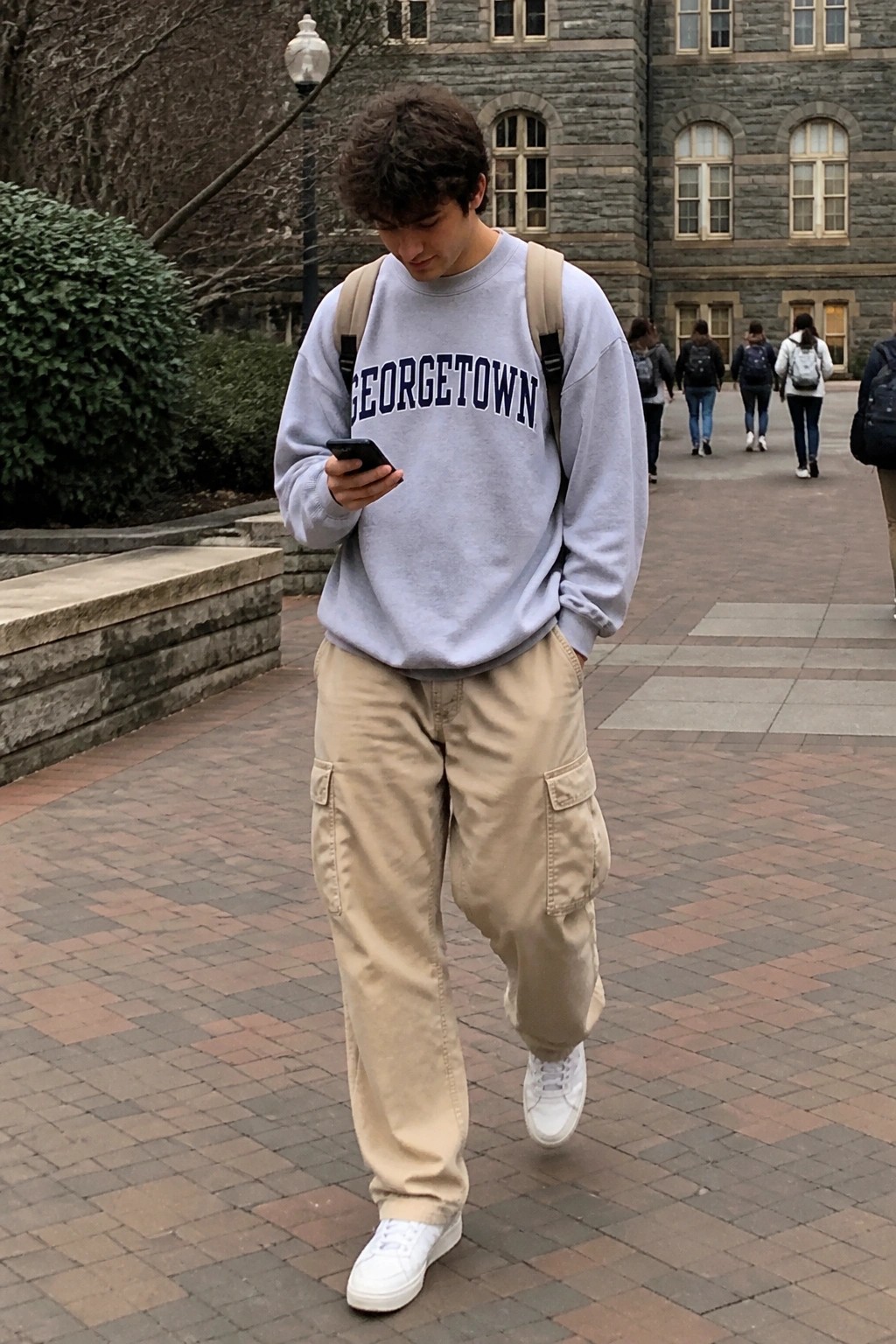 Man wearing light gray Georgetown sweatshirt, beige baggy cargo pants, white sneakers and beige backpack while checking phone on brick walkway.