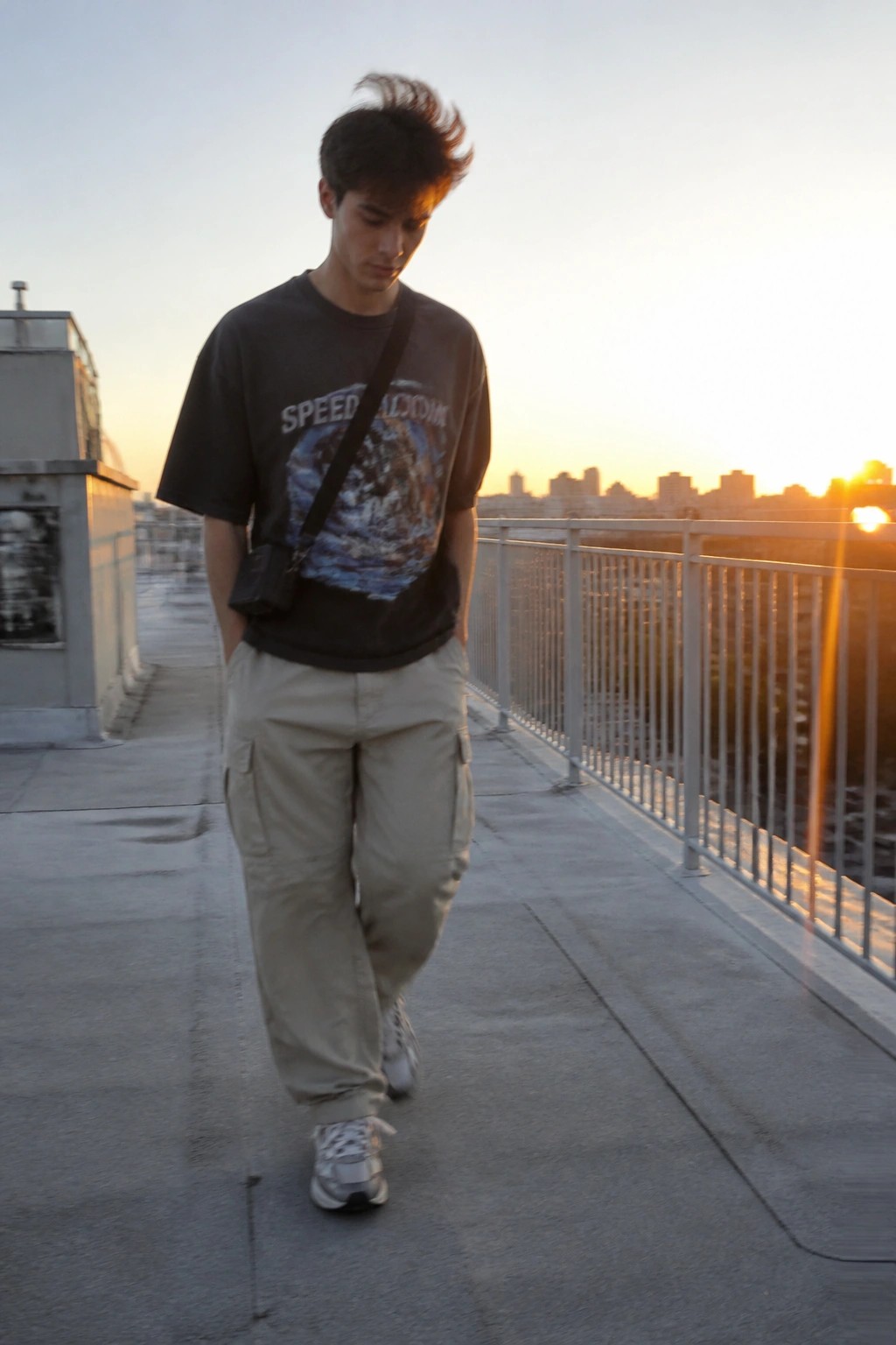 Young man in graphic tee and beige cargo pants walking on rooftop at sunset