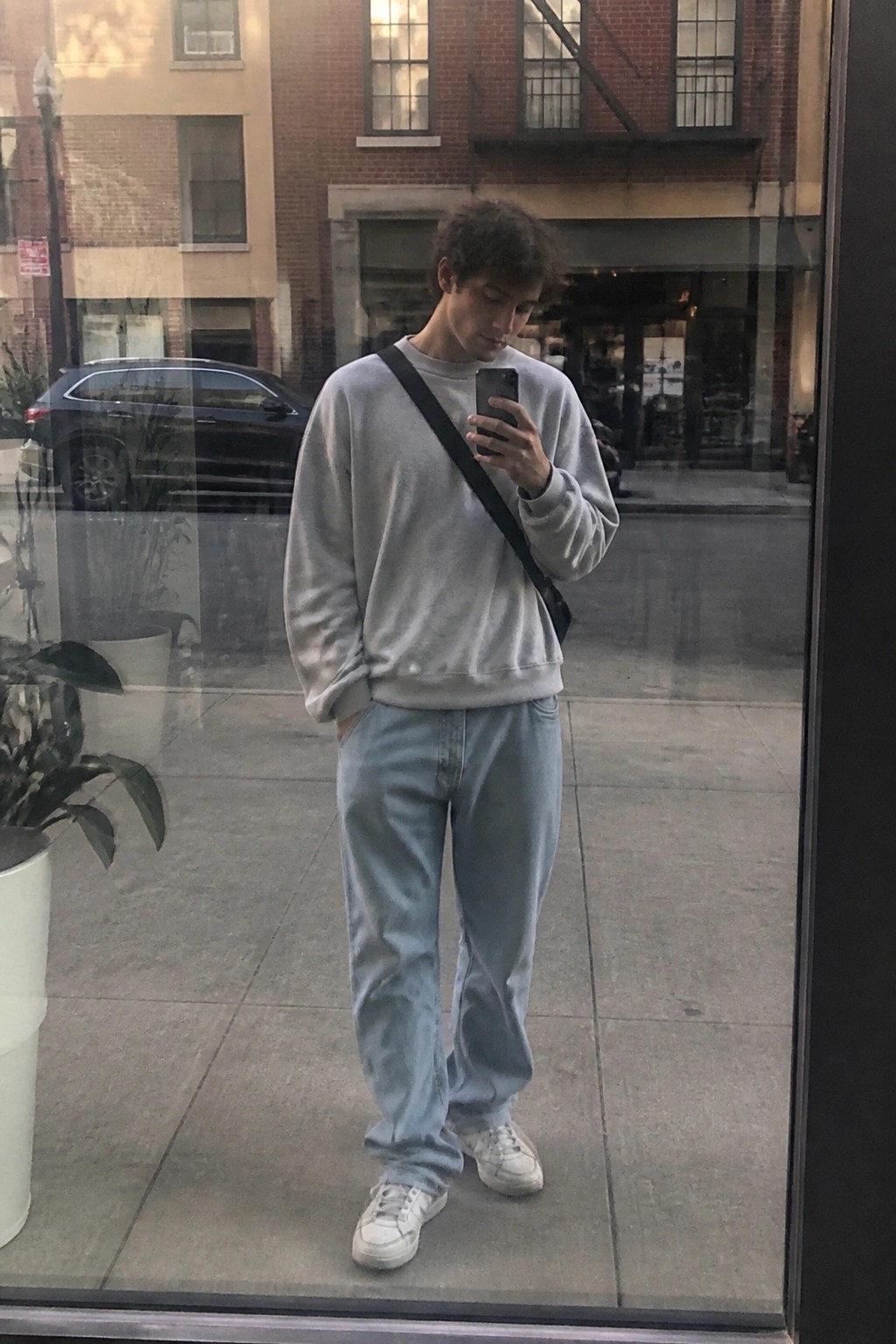 Man wearing light gray sweatshirt, light blue baggy jeans, white sneakers and black crossbody bag while holding a phone in an urban setting.