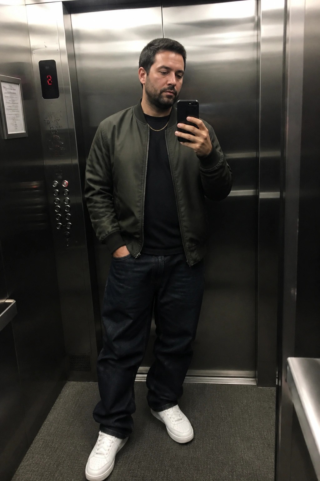 Man in green bomber jacket taking mirror selfie in elevator