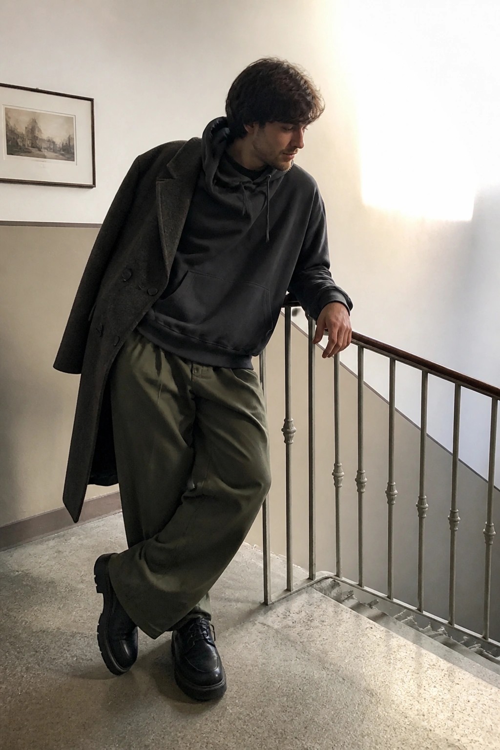 Man in dark overcoat and hoodie with green pants leaning on stair railing