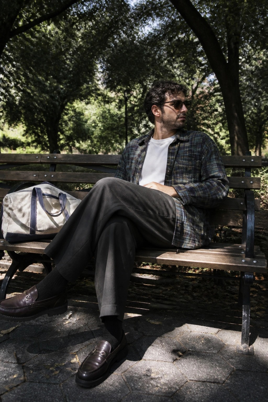 Man in plaid shirt and sunglasses sits on park bench beside white duffel bag